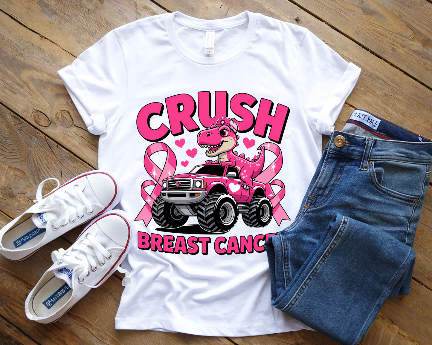 White t-shirt with pink breast cancer awareness design, paired with blue jeans and white sneakers on a wooden surface.